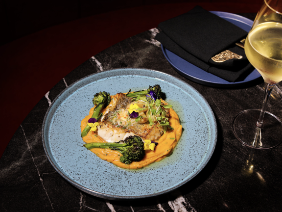 Blue plate with orange coloured puree, green broccoli and fish dish. Gilled glass of white wine stands next to the plate.