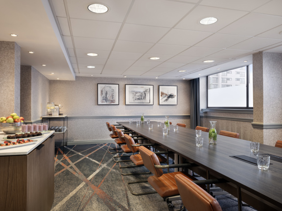 Bright and modern meeting room with large rectangular table surrounded by brown leather dining chairs. To the left side of the room there are table set up with snacks, water and fruit. On the right there is one large window.
