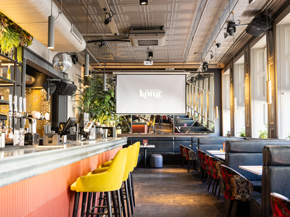 Interior of Kong bar in Glasgow featuring a industrial, modern space with banquet seating and yellow bar stools in front of a peach bar. Daylight streams in through the large windows, hanging greenery, and a well-stocked bar. Large tv screen at the far end reads Kong. 