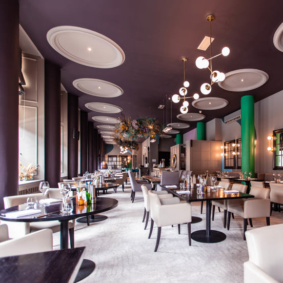 An image of the modern, bright dining room at Glaschu. A long large room with a dark, painted ceiling and cream carpets; plasterwork details on the ceiling and around the large windows in the left wall are painted white. 3 large, cylindrical pillars against the right wall are painted a bright, grassy green. The room is furnished with cream, leather chairs and glassy black tables. Modern spherical light features and a large dried-flower arrangement are suspended from the long ceiling at intervals.