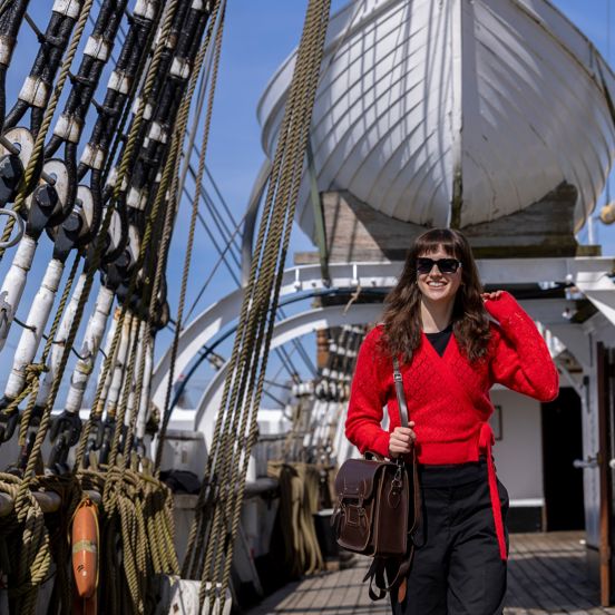 Lady wearing red jumper and sunglasses walks along the deck of The Tall Ship, rigging can be seen on the left hand side with lifeboat raised up in the background of the image