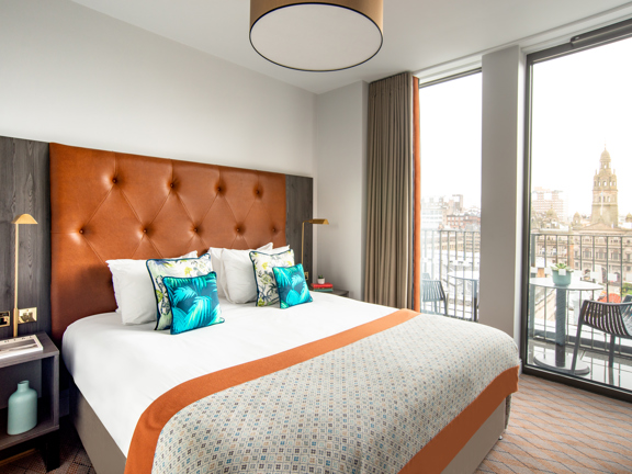 An interior view of a Native Apartment bedroom shows a bright room. The floor is carpeted with a pale grey and orange zig-zag carpet. The bed has a large tan-leather headboard, it is made with crisp white sheets with decorative pillows and a throw in patterns of blue and orange. The far wall is glass and a small, railed balcony is visible outside, with a table and chairs overlooking a steeple and cityscape. The room is decorated and equipped with plugs, lamps, books and potted plants, making it homely.