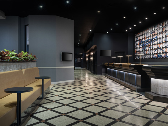 Large hotel lobby and reception. Black and white checked marble floor. On the right is a long reception desk behind which is orange and blue contemporary art work. On the left is a long yellow sofa.