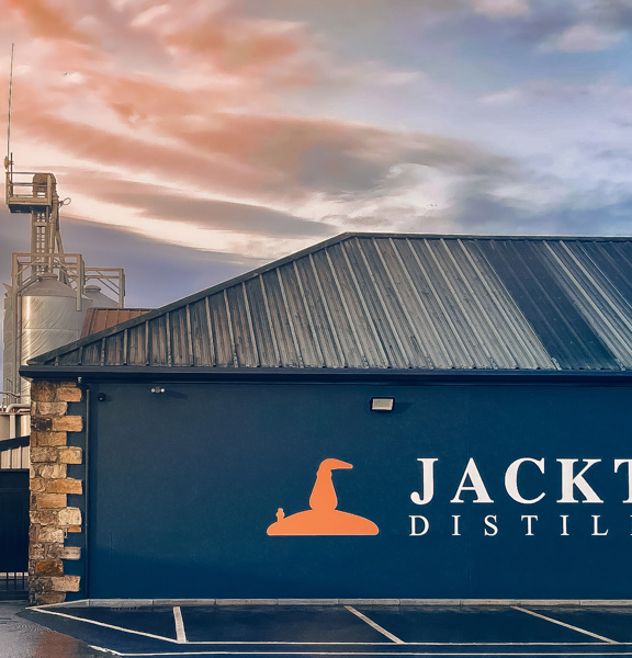 Exterior view of Jackton Distillery at sunset, featuring a dark blue building with the distillery's name and orange logo on the side. Industrial equipment and metal silos are visible in the background, under a colourful sky with dramatic clouds.