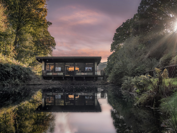 A pool of water sits in front of a wooden building, the building is surrounded by trees on either side. 