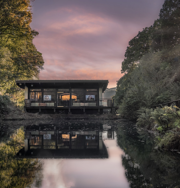 A pool of water sits in front of a wooden building, the building is surrounded by trees on either side. 
