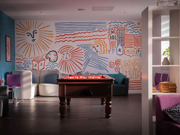 A light and bright social space with a pool table in the middle of the room. Occasional seating and bookshelves are dotted around the space. The room is brightened by blue, yellow and red murals of buildings and faces painted on the far away wall. 