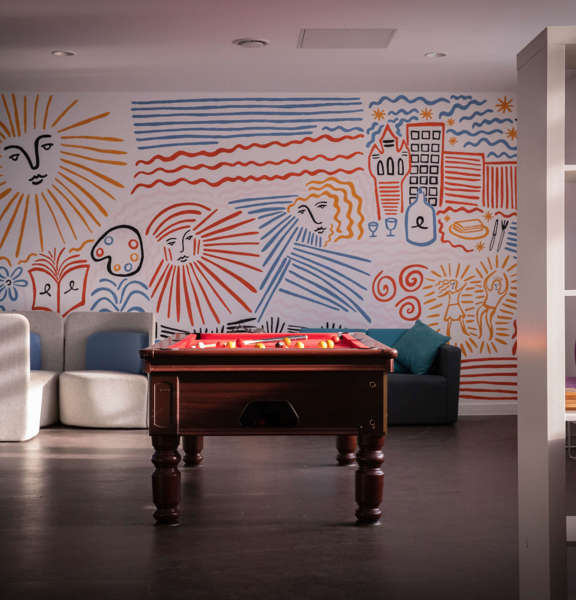 A light and bright social space with a pool table in the middle of the room. Occasional seating and bookshelves are dotted around the space. The room is brightened by blue, yellow and red murals of buildings and faces painted on the far away wall. 