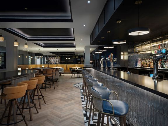 A hotel bar. To the right there is a long black bar top with blue/grey subway tiles along its length. In front of the bar are several blue leather bar stools. To the left hand side are various tables and chairs and at the far end there is a large orange leather banquette. 