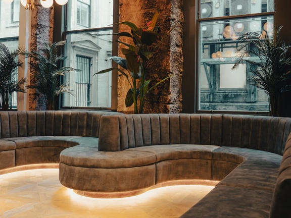 Long and winding brown suede sofa sits below floor to ceiling windows. Large green plants sit behind the sofa which is illuminated from below by a glow of light.