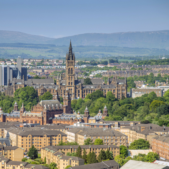 Sunny cityscape with the Gothic revival-style spire of the University of Glasgow's main building, surrounded by greenery and buildings of the West End, including the Baroque-style Kelvingrove Art Gallery and Museum