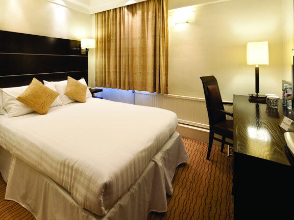 An internal view of a standard, double Mercure hotel bedroom. A small, brightly lit room with white walls, an orange, lined carpet and a curtained window. The bed is on the left hand side, it has a dark wood headboard and is made with white bedsheets. On the right a dark wood desk or dresser can be seen. A lamp, bottle of water and a large TV and its remote are on top and a dark wood, high-backed chair is tucked underneath.