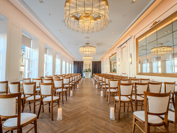 Long bright room set up theatre style - floor to ceiling windows, wooden herringbone flooring white walls with large mirrors and chandeliers hang above. 