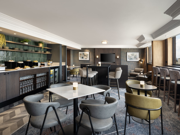 Modern lounge room with various tables and chairs. On the left there is a range of glass fronted fridges filled with drinks and on top a black marble worktop laid with pastries, plates and cakes. 