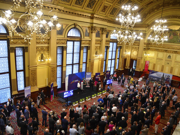 Image of reception taking place in Glasgow City Chambers Banqueting Hall. A large group of people are gathered in a large ornate room with large, arched windows, chandeliers and decorative ceiling. A presentation is taking place on a small stage area, where 4 people are standing in front of banners with a lectern