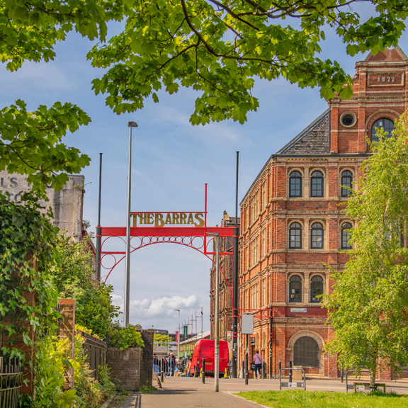 View towards The Barras entrance in Glasgow’s East End, framed by trees and historic red sandstone buildings