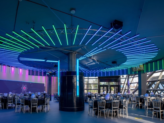 Circular room glazed on left hand side with floor to ceiling windows. Round table set up below a dark ceiling with neon blue and green lights illuminating the room. 