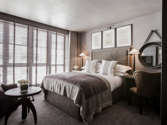 An interior view of a Dakota Deluxe classic bedroom. The bed has a grey, woven blanket over it and grey, velvet headboard. The window has pale curtains and blinds and takes up the entire, furthest wall. There is an armchair and coffee table at the foot of the bed and to the right of the bed a desk, mirror and wing-backed chair can be seen. The room is further decorated with a vase of flowers, framed prints and table lamps. 