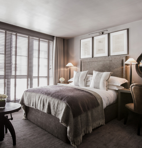 An interior view of a Dakota Deluxe classic bedroom. The bed has a grey, woven blanket over it and grey, velvet headboard. The window has pale curtains and blinds and takes up the entire, furthest wall. There is an armchair and coffee table at the foot of the bed and to the right of the bed a desk, mirror and wing-backed chair can be seen. The room is further decorated with a vase of flowers, framed prints and table lamps. 