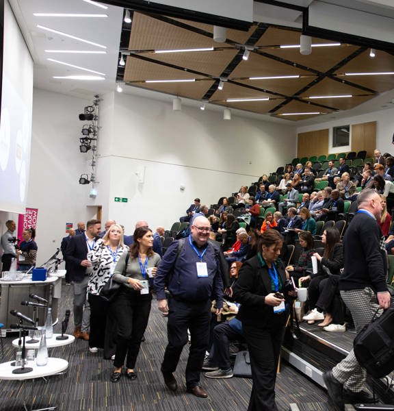 A large attendance of men and women, some standing some sitting down, in a big room