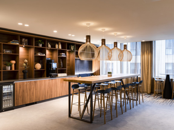 Large breakout space with wooden units and shelves running along one wall. High table and bar stools running down middle of room. 
