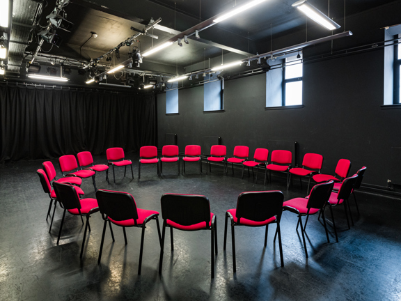 An all black room with red chairs set up in a circle, white strip lights hang from the ceiling and four windows let in diffused light. 