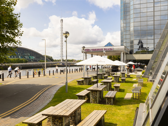 Picnic benches below white parasols that read Crowne Plaza facing onto the river