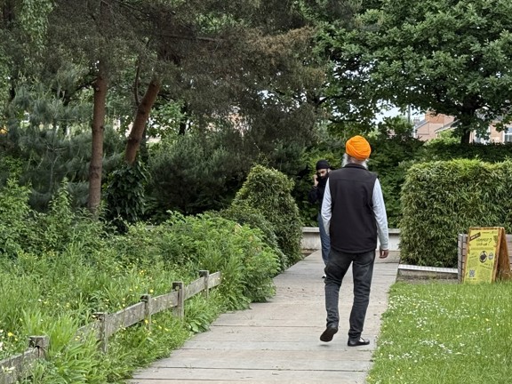 Man walking along garden path with grass and shrubs at either side and trees in the background