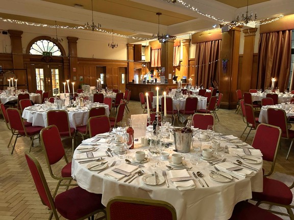 Large room with parquet flooring and wooden panelled walls. Fairy lights hand from the ceiling and the round tables are set with crisp white linen tablecloths, silver cutlery, glasses and candles.