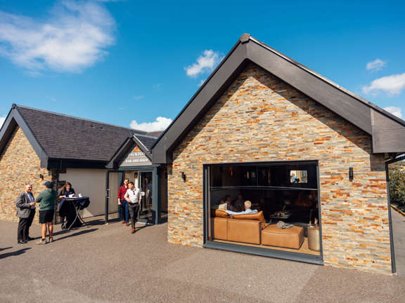 Jackton Distillery bar and shop is housed within a modern building with decorative stonework on to the front. People can be seen coming and going from the distillery. 