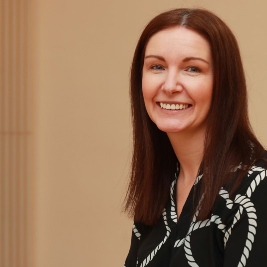 Kay Morrison wearing a black shirt with white pattern smiling at the camera with a beige background