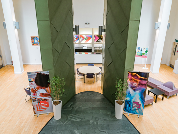 Large white room with double height ceilings and wooden flooring.  Two large green pillars sit side by side in the room surrounded by occasional seating. 