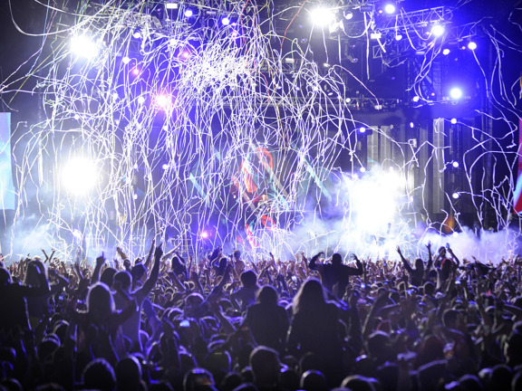 A cheering crowd is silhouetted in front of stage at Radio 1's Big Weekend Glasgow, the lights are blue and string confetti is falling from the sky.