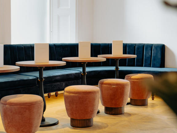 Long dark velvet banquette faces four round wooden tables sat opposite four tan leather stools. 