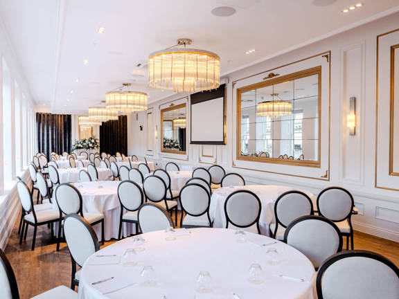 Long bright white room with three large chandeliers. Round tables set with white linen surrounded by dark wooden framed chairs with white seat and back cushions. Large mirrors line the right hand side of the room opposite large windows.
