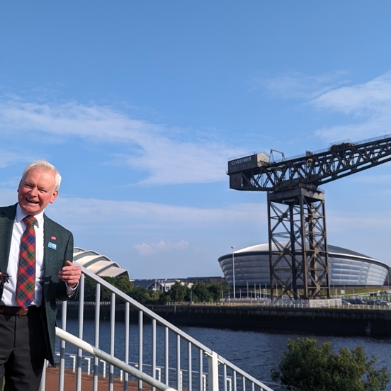 Tour guide standing in the sunshine on the bank of the River Clyde, Finnieston crane and SEC & Hydro.