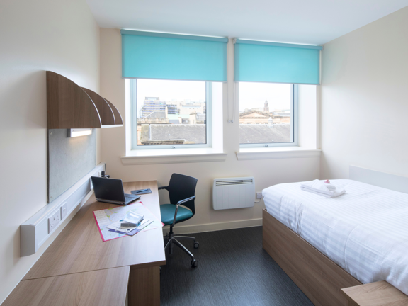 Bright bedroom with single bed, wooden desk with matching shelves above. A pale blue roller blind frames the window. 