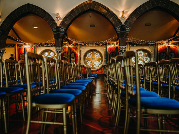 An interior image from Oran Mor taken across the width of an event space, & the former parish church. The aisle is separated from the main nave by a series of pointed arches with carved stone pillars. Fairy lights are draped from the ceiling and rich wall paintings and circular stained-glass windows punctuate the far, outer wall. The floor is a highly polished wood and its width is covered with rows of gold chairs with blue velvet seats. 