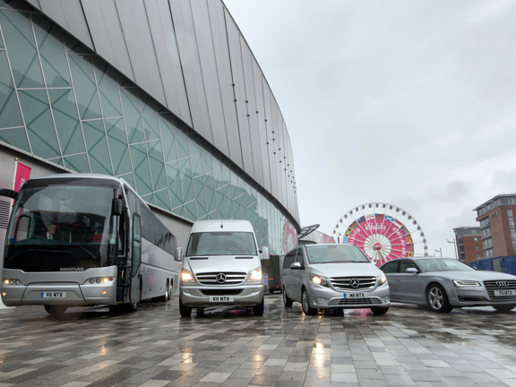 A silver coach, van, people carrier and car are parked in a row besides the silver, curving wall of a large modern building. The ground is paved with stone slabs, wet from rain. The sky is grey and the cars headlights are on. A white ferriswheel with pink branding, blue boarding and 2 modern, brick buildings can all be see in the background.