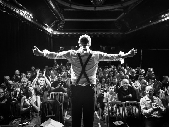 Man on stage with his arms outstretched facing a smiling and clapping audience. 