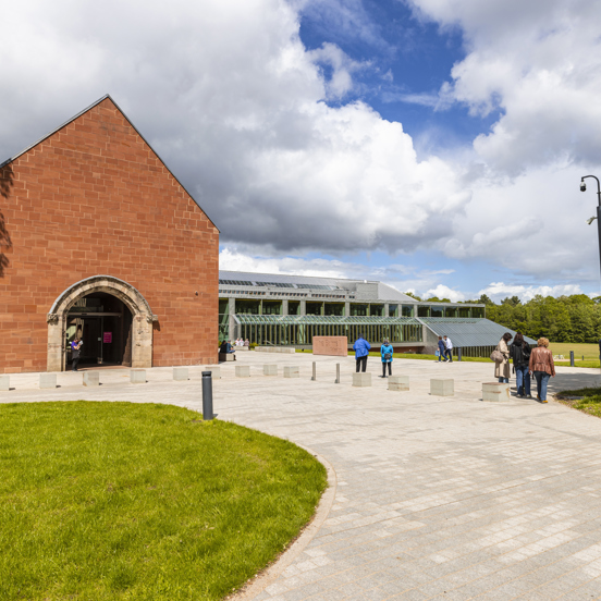 An external view of the Burrell Collection. A paved walkway leads up to the red sandstone entrance of the museum. A large glass building and green lawn is visible against trees and a cloudy sky.