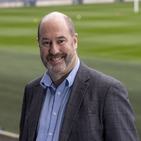 A photograph of Commonwealth Games Scotland Chief Executive Jon Doig OBE with a green pitch in the background
