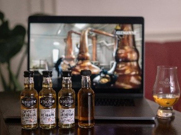 Four miniature bottles of whisky each with a Glengoyne label on them. A glass with whisky in it sits to the side and behind is a picture of copper stills in a distillery