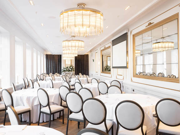 Long bright white room with three large chandeliers. Round tables set with white linen surrounded by dark wooden framed chairs with white seat and back cushions. Large mirrors line the right hand side of the room opposite large windows.