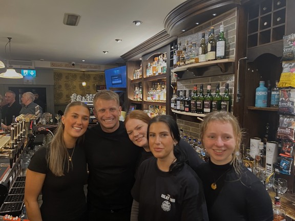 A group of five smiling bar staff members posing together behind a well-stocked bar in a warmly lit pub. Shelves of various whisky bottles and snacks are displayed in the background, along with rustic decor and signage that reads "Slàinte Mhath - Good Health!". Other patrons are visible in the background, engaged in conversation.