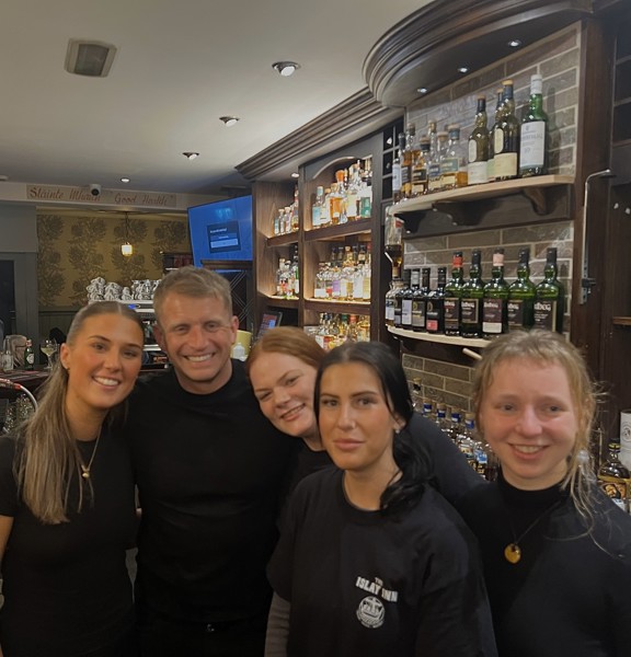 A group of five smiling bar staff members posing together behind a well-stocked bar in a warmly lit pub. Shelves of various whisky bottles and snacks are displayed in the background, along with rustic decor and signage that reads "Slàinte Mhath - Good Health!". Other patrons are visible in the background, engaged in conversation.