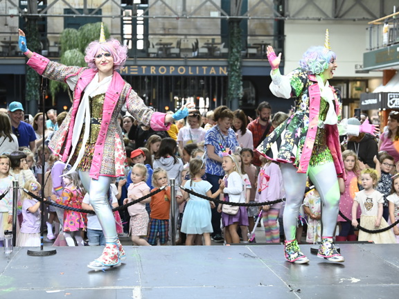 Two performers wearing unicorn horns and colourful costumes dance on stage in front of a crowd of children in Merchant Square.