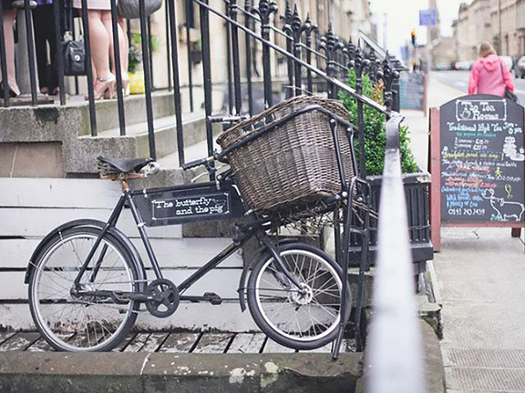 An exterior detail shot of The Butterfly and The Pig restaurant. 6 stone steps lead up from a wide tarmac pavement. A blackboard A-frame sign with colourful chalk writing sits at the base of the stairs. Black wrought iron railings line either side of the stairs. A black bicycle leans against the side of the staircase; it has a large wicker basket over the front wheel and is decorated with a small sign advertising the restaurant. The feet of a party of guests can be seen on the top step, wearing heels.