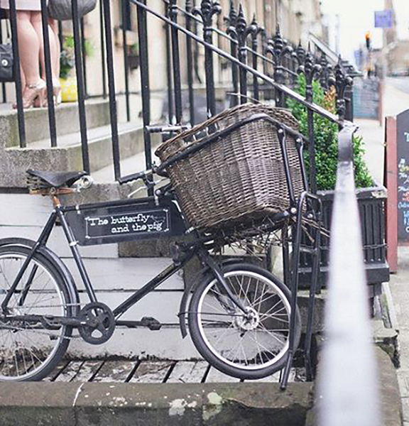 An exterior detail shot of The Butterfly and The Pig restaurant. 6 stone steps lead up from a wide tarmac pavement. A blackboard A-frame sign with colourful chalk writing sits at the base of the stairs. Black wrought iron railings line either side of the stairs. A black bicycle leans against the side of the staircase; it has a large wicker basket over the front wheel and is decorated with a small sign advertising the restaurant. The feet of a party of guests can be seen on the top step, wearing heels.