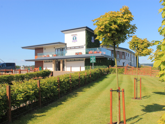 Modern white building sits at the end of a drive surrounded by countryside and trees